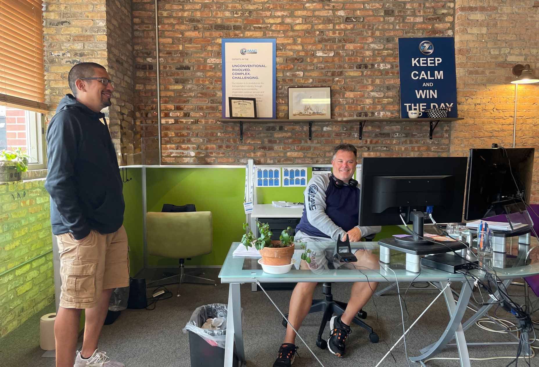 Two members of the ZMac team in our office. One sits at his computer while the other stands next to him.